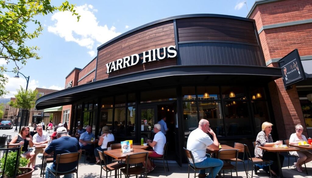 A vibrant street view showcasing a Yard House restaurant location during a sunny day. In the foreground, a well-maintained outdoor patio with stylish dining sets, where patrons in casual clothing enjoy their meals and drinks, smiles on their faces. The middle ground features the distinctive dark wood and glass facade of the Yard House, with its recognizable logo above the entrance and large windows displaying the bustling interior, filled with lively conversations. In the background, a clear blue sky with a few fluffy clouds and a hint of urban scenery, such as trees and nearby storefronts, emphasizing a lively neighborhood atmosphere. Bright, natural lighting illuminates the scene, creating a warm and inviting ambiance, captured with a wide-angle lens to highlight the restaurant's inviting exterior.